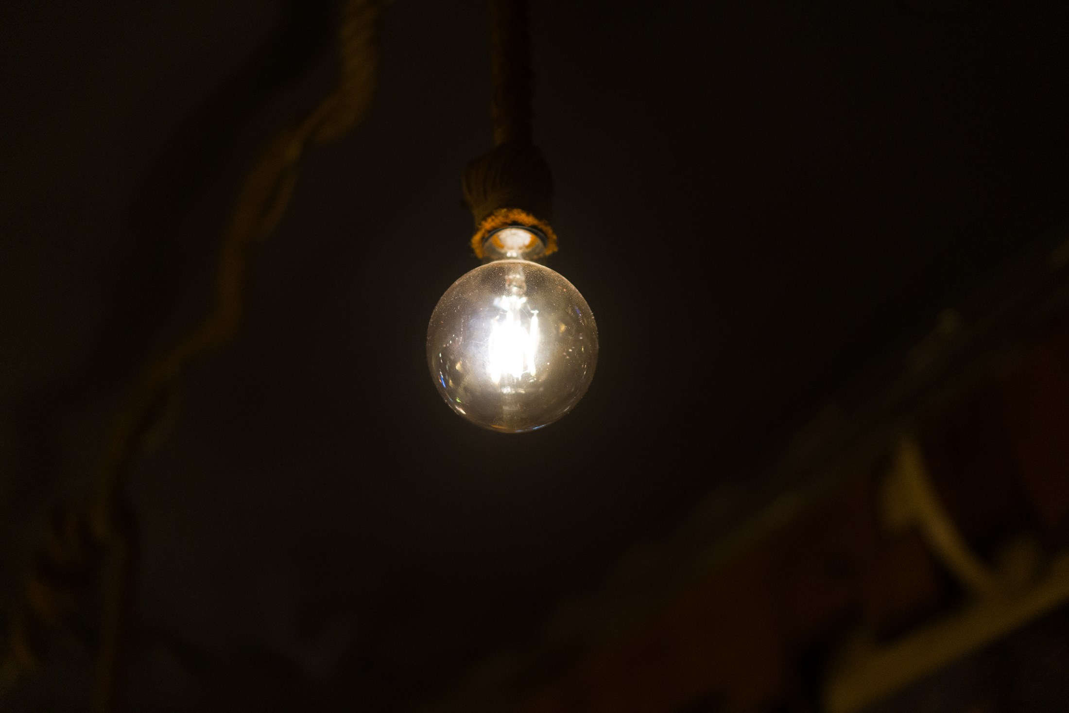 A single bare incandescent bulb hanging from a rope in a dark room, its filament burning warm orange against the black.