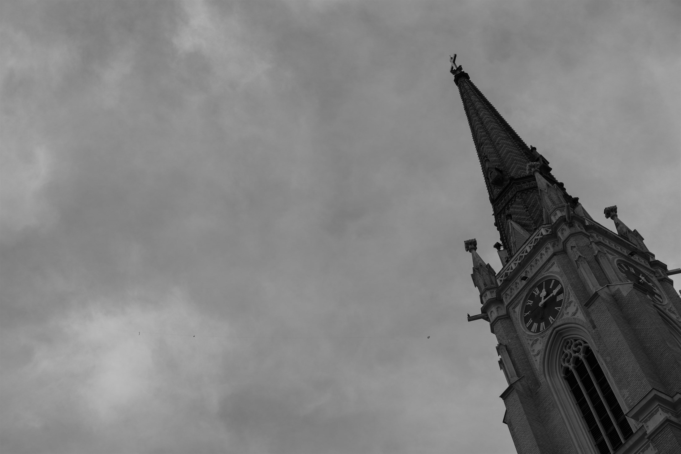 A church spire in black and white seen from below, leaning into a sky of heavy overcast, its clock face and stone finials picked out against the cloud.