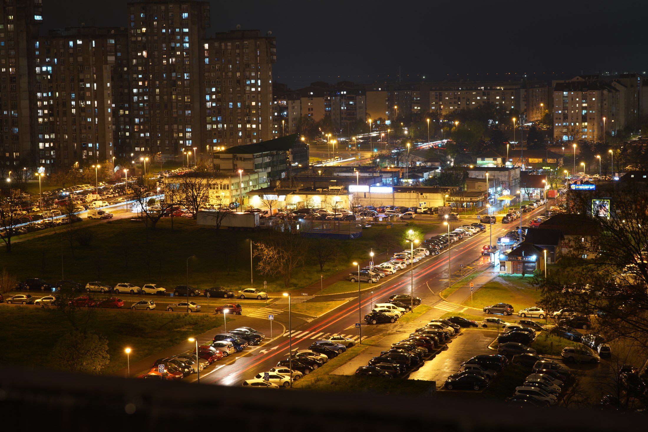 A city from above at night on a long exposure, tower blocks lit window by window and a boulevard of traffic running out to the far edge.