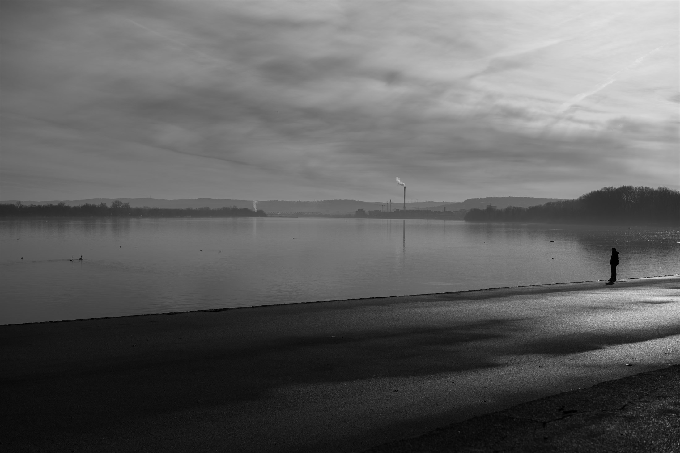 A wide lake at first light in black and white, one figure standing alone on the wet shore, mist lying along the far bank.