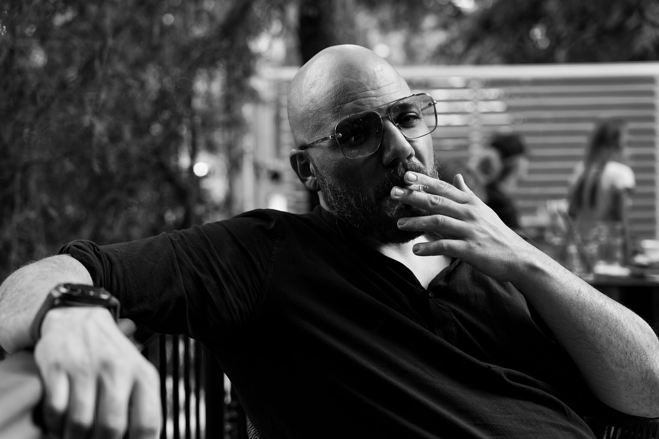A bearded man in aviator sunglasses sitting back on a café terrace in black and white, one arm along the chair, a hand raised to his mouth.
