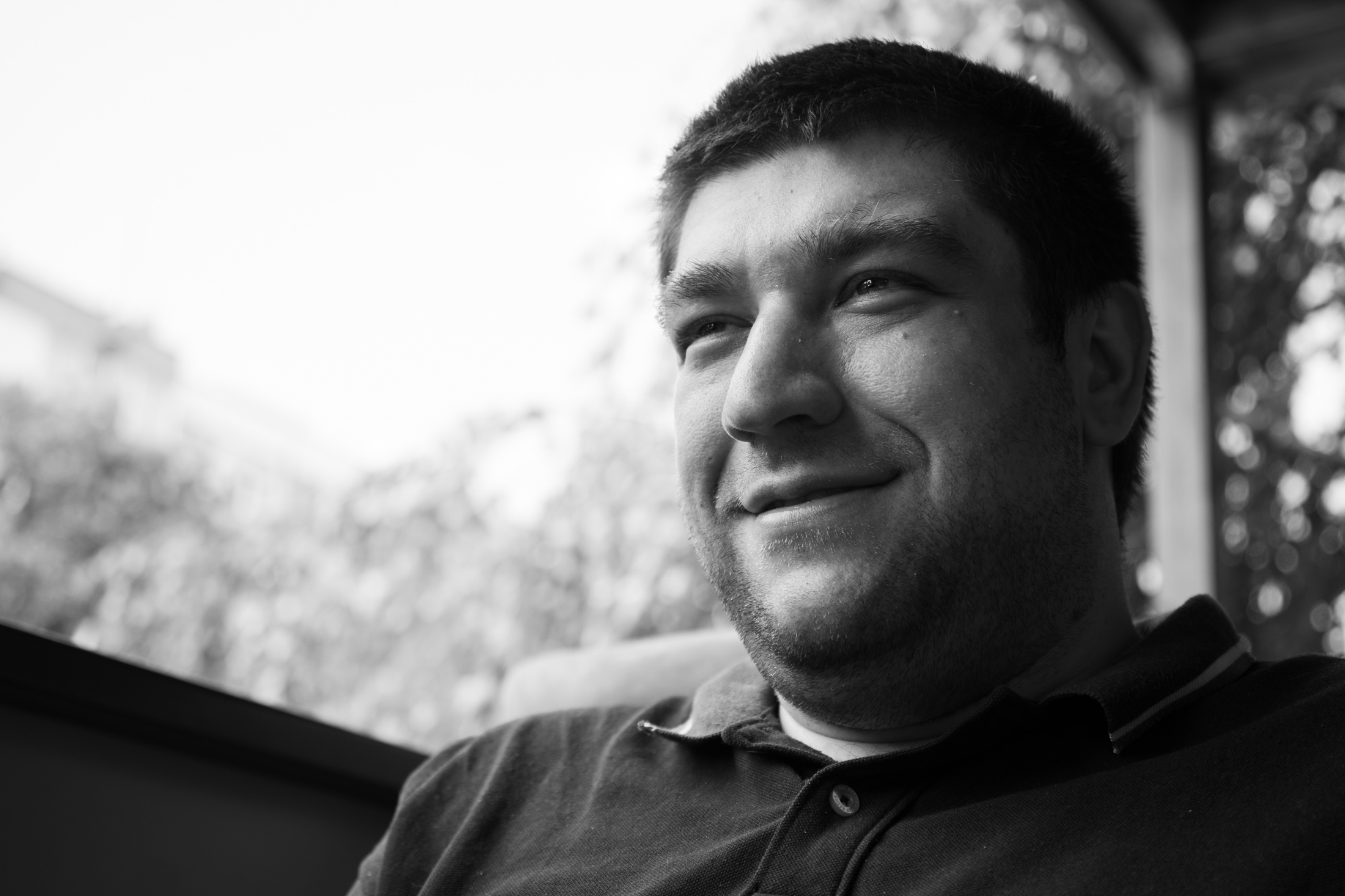 A man on a terrace in black and white, half-smiling, looking off past the camera into bright afternoon light.